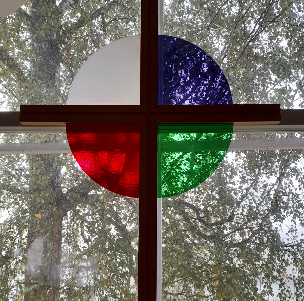 Kuvassa ikkunaa vasten oleva risti, jonka keskellä ympyrä, missä valkoinen, punainen, vihreä ja sininen väri